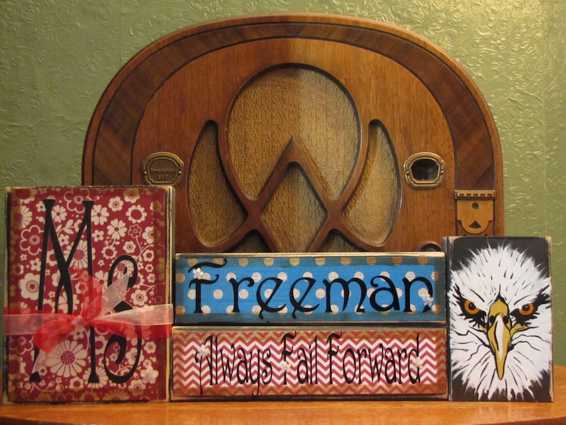 May include: Three wooden blocks with painted designs. The first block has a red and white floral pattern with the letter "M" in black. The second block is blue with white polka dots and the word "Freeman" in black. The third block has a red and white chevron pattern with the words "Always Fall Forward" in black. The last block has a black background with a white and yellow eagle head.