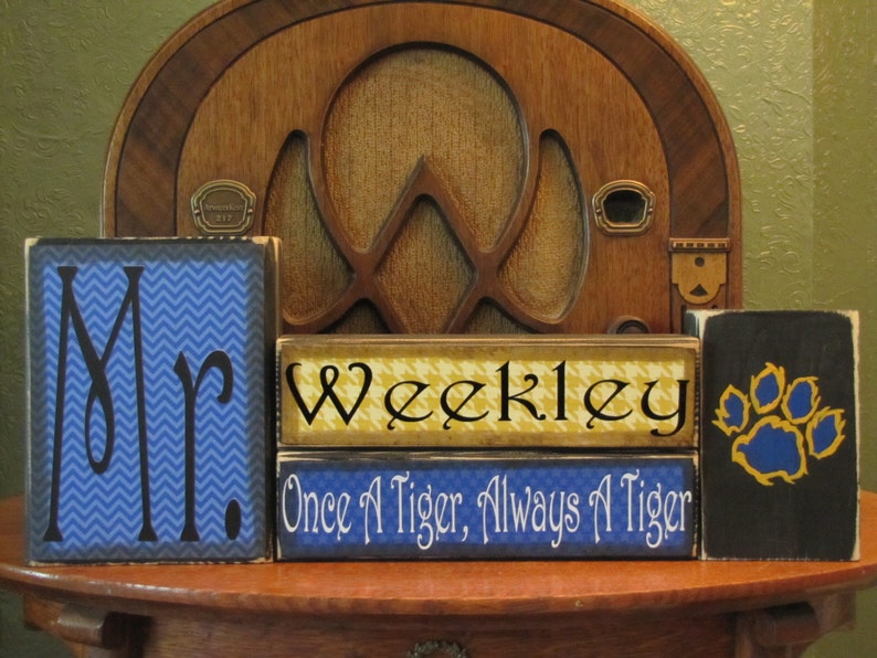 May include: Three wooden blocks with blue and yellow paint. The first block says "Mr." in black lettering on a blue chevron pattern. The second block says "Weekley" in black lettering on a yellow and black checkered pattern. The third block says "Once A Tiger, Always A Tiger" in white lettering on a blue background. The last block has a black background with a yellow paw print with blue claws.