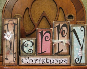 May include: Wooden blocks spelling out "Merry Christmas" with decorative paper and embellishments. The blocks are painted in various colors, including pink, blue, and brown.