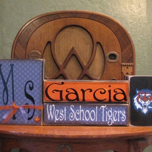 May include: Three wooden blocks stacked on a table. The top block is black with a blue and orange tiger graphic. The middle block is orange with the text "Garcia" in black. The bottom block is blue with white polka dots and the text "Ms" in black. The bottom block also has a small orange bow and a small orange flower.