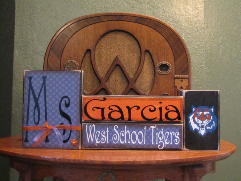 May include: Three wooden blocks stacked on a table. The top block is black with a blue and orange tiger graphic. The middle block is orange with the text "Garcia" in black. The bottom block is blue with white polka dots and the text "Ms" in black. The bottom block also has a small orange bow and a small orange flower.