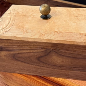 Könnte beinhalten: Eine Holzkiste mit einem Scharnierdeckel und einem Messingknopf. Die Oberseite der Kiste ist aus Ahornholz mit einer hellbraunen Oberfläche. Die Unterseite der Kiste ist aus Nussbaumholz mit einer dunkelbraunen Oberfläche.