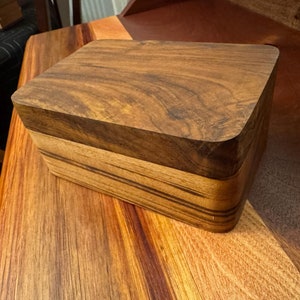 May include: A wooden box with a dark brown lid and a lighter brown base. The box has a smooth, polished finish and is made of high-quality wood.