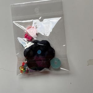 Könnte beinhalten: Eine transparente Plastiktüte mit verschiedenen Bastelmaterialien. Enthält ein schwarzes, pfotenformiges Filzornament mit blauem und gelbem Design, eine rosa Schleife, eine rosa Blume, einen hellblauen Kreis und bunte Pailletten.