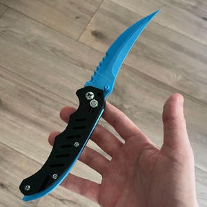 Pode incluir: Uma faca dobrável preta e azul com uma lâmina azul curva e serrilhada. A pega é preta com um detalhe azul. A faca é segurada em uma mão.