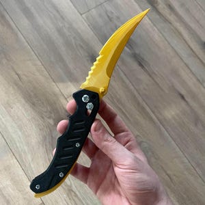 Könnte beinhalten: Ein Klappmesser mit einer gebogenen, gelben Klinge und einem schwarzen Griff. Die Klinge hat eine gezahnte Kante in der Nähe des Griffs. Der Griff ist schwarz mit einem strukturierten Design und mit silbernen Schrauben befestigt. Das Messer ist geöffnet.