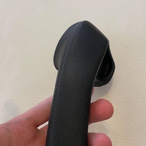 May include: A black, textured, curved handle with a matte finish. The object features a solid, elongated grip section that tapers slightly toward the top, with a wider, rounded base component visible at the upper end of the structure.