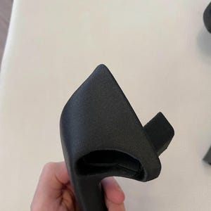 May include: A matte black, textured, 3D-printed plastic handle component with a curved, ergonomic shape. The object features a hollow interior section and a protruding rectangular tab on one side, showcasing a granular surface finish throughout.