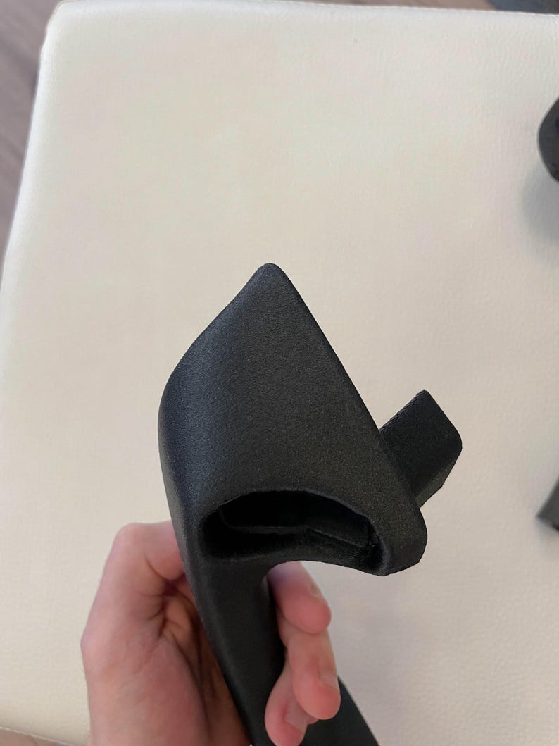 May include: A matte black, textured, 3D-printed plastic handle component with a curved, ergonomic shape. The object features a hollow interior section and a protruding rectangular tab on one side, showcasing a granular surface finish throughout.