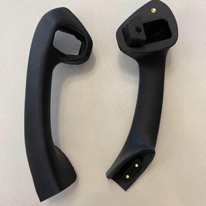 May include: Two black textured automotive grab handles with contoured shapes. Each handle features integrated mounting points with small metallic gold-colored contact pins visible at the connection ends, designed for secure installation within a vehicle interior.