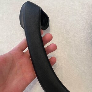 May include: A matte black, curved automotive interior door handle with a textured finish. The handle features a rounded, ergonomic grip section that tapers toward the bottom and includes a molded mounting housing at the top end.