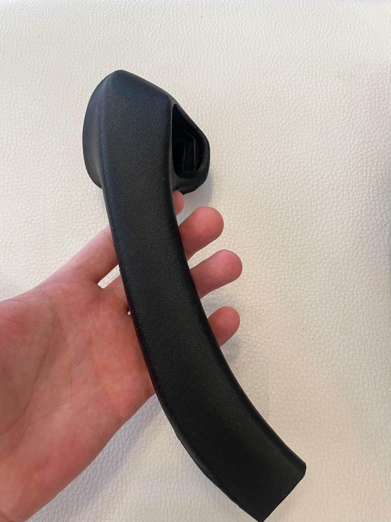 May include: A matte black, curved automotive interior door handle with a textured finish. The handle features a rounded, ergonomic grip section that tapers toward the bottom and includes a molded mounting housing at the top end.