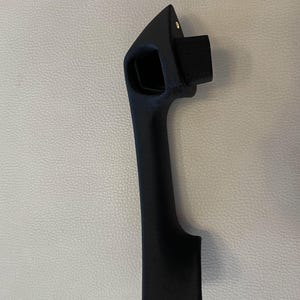 May include: A matte black automotive door handle with a textured finish. The handle features an elongated vertical grip, a rectangular mounting protrusion at the top, and a small metallic screw visible near the upper edge of the handle assembly.