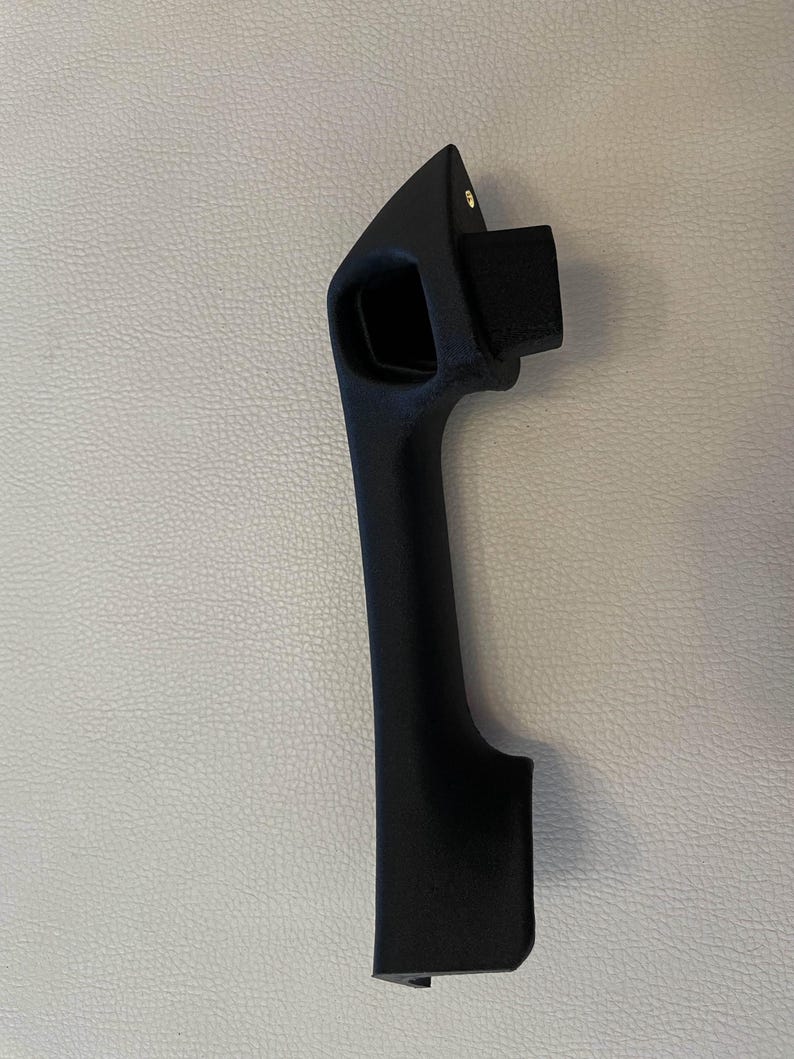 May include: A matte black automotive door handle with a textured finish. The handle features an elongated vertical grip, a rectangular mounting protrusion at the top, and a small metallic screw visible near the upper edge of the handle assembly.