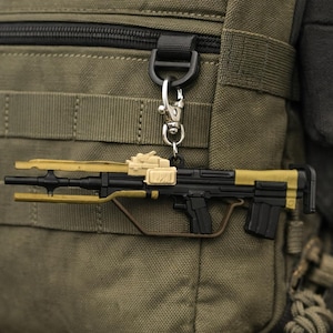 May include: A miniature black and tan rifle keychain with a silver clasp, attached to a green backpack. The keychain features a detailed design, including a barrel, stock, and trigger guard. The backpack has multiple straps and pockets.
