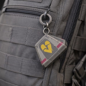 May include: A gray, diamond-shaped keychain with a yellow broken heart design and pink accents. The keychain is attached to a silver ring and is hanging on a gray backpack.