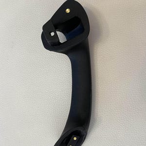 May include: A matte black automotive grab handle featuring a curved, ergonomic design. The handle includes a top mounting point with a single brass-colored metal insert and a bottom mounting point with two brass-colored metal inserts for secure attachment.