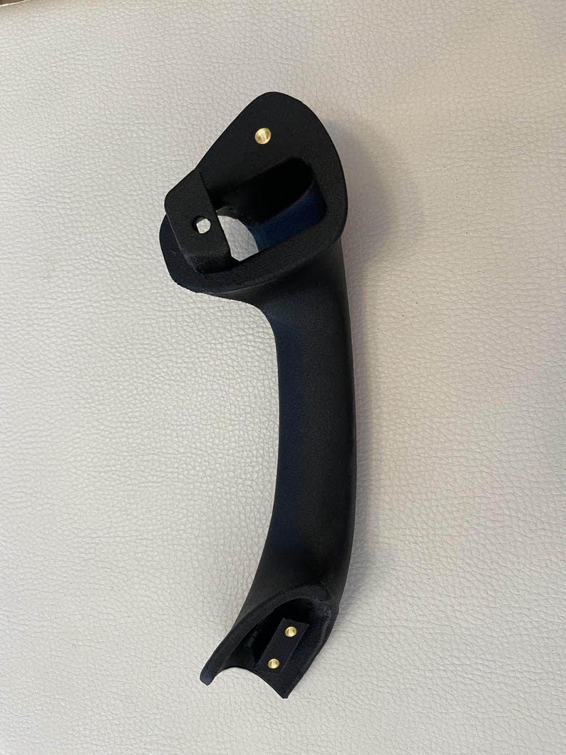 May include: A matte black automotive grab handle featuring a curved, ergonomic design. The handle includes a top mounting point with a single brass-colored metal insert and a bottom mounting point with two brass-colored metal inserts for secure attachment.