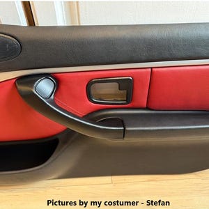 May include: A car door panel featuring a black upper and lower section with a central red leather insert. The panel includes a black door handle, a black interior door release lever, and a speaker grille. Text at the bottom reads: Pictures by my costumer - Stefan