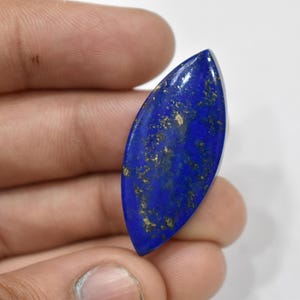 May include: A polished, elongated lapis lazuli gemstone. The deep blue stone has gold flecks and a smooth, glossy surface. The gemstone is held in a hand against a white background. This gemstone is ideal for jewelry making.