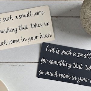 May include: Two rectangular signs with the text "Cat is such a small word for something that takes up so much room in your heart." One sign is off-white, the other is dark gray. The signs are on a white wooden surface.