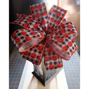 May include: A large Halloween bow featuring a black and white checkered pattern with orange jack-o'-lanterns. The bow is made of ribbon and is likely used for decoration. The bow is placed on top of a black and silver lantern.