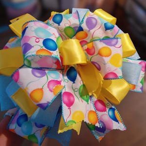 May include: A large, decorative bow featuring a white ribbon printed with colorful balloons. The bow has yellow and light blue satin ribbons, creating a layered effect. The edges of the ribbon are trimmed with a blue and white stitched border.
