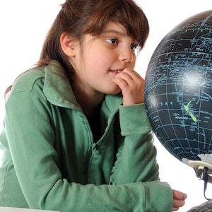 Può includere: Una giovane persona con una giacca verde con cerniera guarda un mappamondo nero su un supporto metallico. Il testo "CHE COS'E?" e "LA GEOGRAFIA?" è visibile sopra la testa della persona.