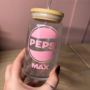 May include: A clear glass tumbler with a wooden lid and a clear straw. The tumbler features a pink circular graphic with the word "PEPSI" and the word "MAX" in white. The tumbler is designed to resemble a soda can.