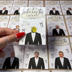 May include: A rectangular game card featuring the text Who has the GROOM? and Scratch to reveal. The card displays a graphic of Shrek in a tuxedo, the handwritten text Fern's Hen Party, and several black bow illustrations on a white background.