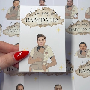 May include: A scratch-off card with the text "Who has the BABY DADDY" in a brown cloud design. The card features a cartoon image of a man holding a baby in a carrier. Other cards with different images are in the background.