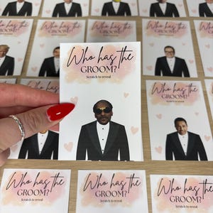 May include: A rectangular game card featuring the text Who has the GROOM? Scratch to reveal in black script and block lettering. The card displays a graphic of a person in a black tuxedo and sunglasses, surrounded by small light pink hearts.