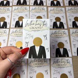 May include: A rectangular white card featuring the text Who has the GROOM? Scratch to reveal and Fern's Hen Party. The card displays a graphic of a tuxedo with a circular gold scratch-off sticker as the head and small black bow illustrations.