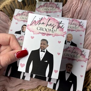 May include: A rectangular game card featuring the text Who has the GROOM? Scratch to reveal in pink watercolour script and black font. The card displays a graphic of a man in a black tuxedo with a bow tie, surrounded by small pink hearts.
