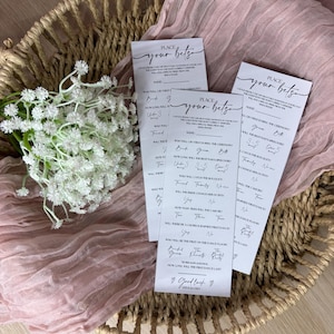 May include: Three white cards with the text "Place Your Bets" are arranged on a woven basket with a pink fabric and white flowers. The cards have fill-in-the-blank questions about a wedding.