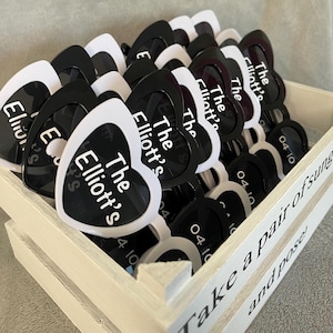Peut inclure: Une collection de lunettes de soleil en forme de cœur dans une caisse en bois blanche. Les lunettes sont noires et blanches, avec le texte "The Elliott's" imprimé sur les verres. La date "04 10" est également imprimée. La caisse a le texte "Take a pair of sunglasses and pose!"