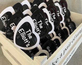 May include: A collection of heart-shaped sunglasses with white frames and black lenses, each printed with the text The Elliott's and 04-10. The sunglasses are stored in a white wooden crate featuring the text Take a pair of sunglasses and pose!