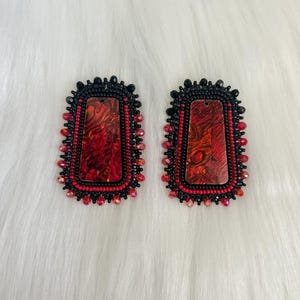 May include: A pair of rectangular earrings featuring a central red and black marbled inlay. The edges are intricately bordered with rows of small black beads and a final outer layer of faceted red beads, creating a textured, beaded frame.