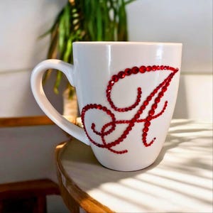 Può includere: Tazza in ceramica bianca con una grande lettera "A" rossa tempestata di gioielli sul davanti. La lettera è composta da piccole pietre rosse rotonde. La tazza ha un manico curvo e poggia su una superficie chiara.