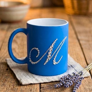 Könnte beinhalten: Eine blaue Keramiktasse mit weißem Innenraum und einem mit Strasssteinen besetzten Henkel. Der Buchstabe "M" ist mit Strasssteinen verziert. Die Tasse steht auf einem grauen Tuch, mit Lavendelzweigen in der Nähe.