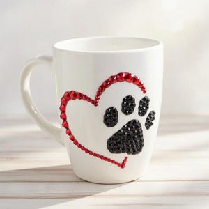 Könnte beinhalten: Weiße Keramiktasse mit Herz- und Pfotenabdruck-Design. Das Herz ist mit roten Strasssteinen umrandet, der Pfotenabdruck ist mit schwarzen Strasssteinen gefüllt. Die Tasse hat einen gebogenen Henkel.