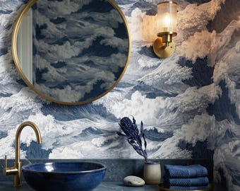 May include: A round mirror with a gold frame hangs above a dark blue vessel sink and a gold faucet. The wall features blue and white wave-patterned wallpaper. A gold wall sconce, a dried flower, and a stack of blue towels are present.