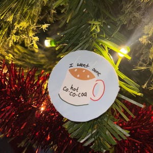 May include: A round, light blue Christmas ornament with the words "I want some hot co-co-coa" and a drawing of a mug of cocoa. The ornament is hanging on a Christmas tree with red tinsel and green needles.