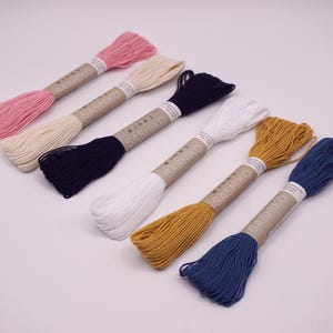 May include: Six spools of sashiko thread in various colors, including pink, cream, navy blue, white, mustard yellow, and denim blue. Each spool is labeled "Sashiko" with a Japanese character.