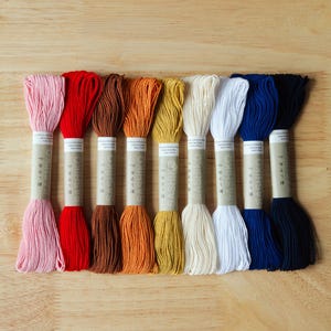 May include: A set of nine skeins of Sashiko embroidery thread in a variety of colors including pink, red, brown, orange, yellow, cream, white, blue, and navy. Each skein is secured with a paper band labeled Sashiko and Made in Japan.