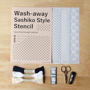 Könnte beinhalten: Ein Wash-away Sashiko Style Schablonen-Set mit einem beigen Anleitungsblatt mit Text, drei weißen Schablonenbögen mit Kreuz- und Wellenmustern, drei Stickgarnsträngen in Marineblau, Weiß und Creme, einer Nadelpackung, einer Schere und einem schwarzen Werkzeug.