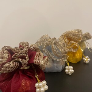 May include: Three small, decorative drawstring bags made of sheer fabric with gold lace trim and pearl accents. The bags are red, yellow, and grey.