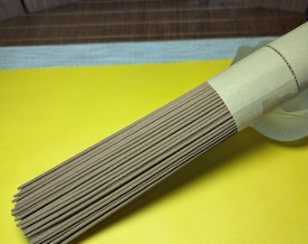 May include: A bundle of incense sticks, wrapped in brown paper, rests on a bright yellow surface. The incense sticks are a light brown color and appear to be neatly arranged. The background includes a decorative screen.