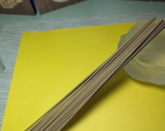 May include: A collection of brown incense sticks, angled on a yellow surface. The incense sticks are slender and cylindrical, with a light brown hue. A decorative screen and a glass dish are visible in the background.