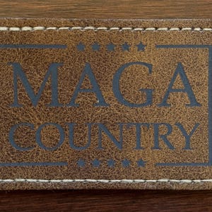 以下が含まれることがあります： 「MAGA COUNTRY」の文字がダークブルーで書かれた長方形の茶色の革パッチ。テキストは、上部と下部に星が付いたダークブルーの長方形の境界線の中にあります。パッチの端には白いステッチが施されています。